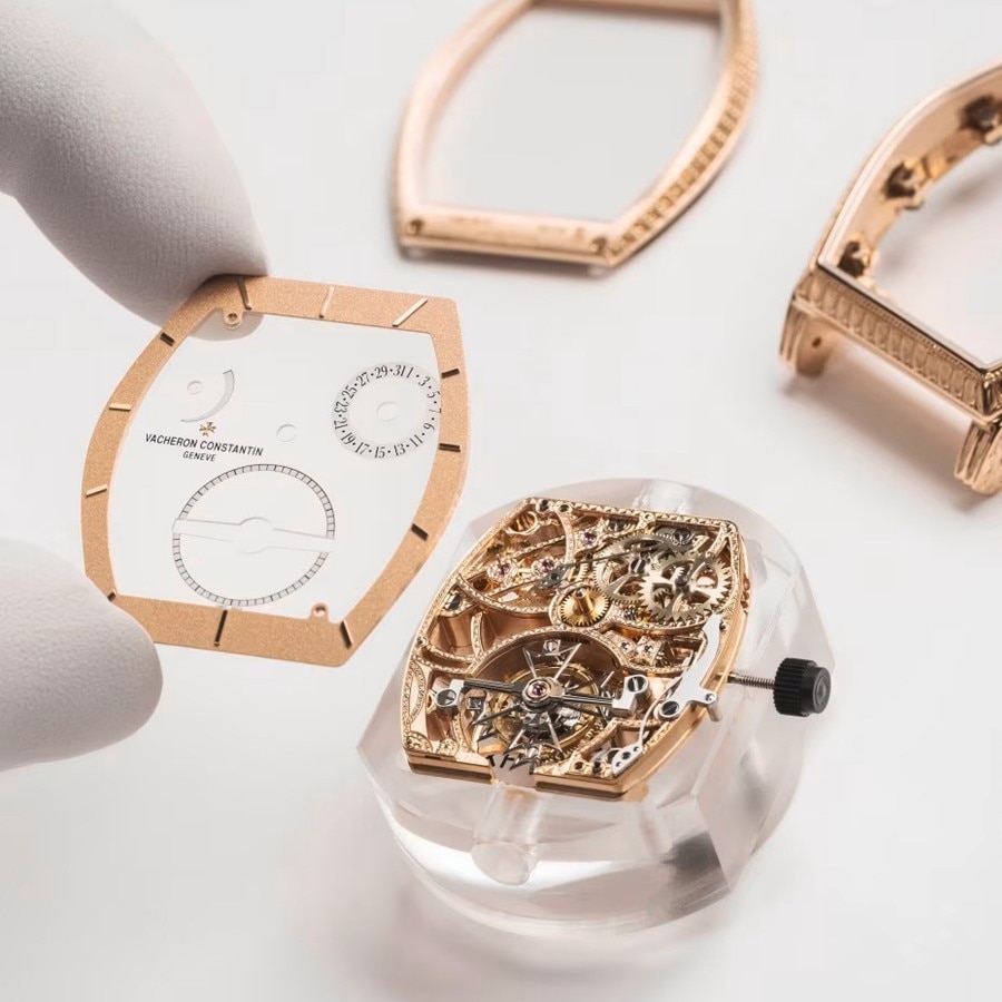 Close-up of a hand in white gloves placing a gold watch bezel on a transparent case with a gold intricate mechanism